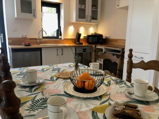 Table de salle à manger avec vaisselle, fruits et cuisine lumineuse en arrière-plan.