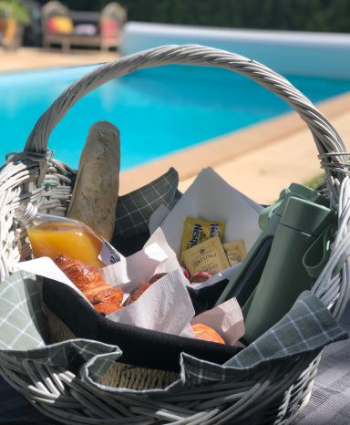 Petit-déjeuner gourmand en bord de piscine – Gîte de charme Les Loges de Bacchus Panier en osier avec des fruits, des boissons et des collations près d'une piscine.