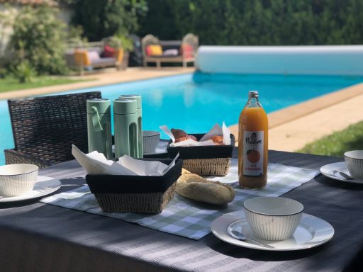 Petit-déjeuner fait maison au bord de la piscine – Les Loges de Bacchus Table mise en place avec des assiettes, boissons et collations près d'une piscine.