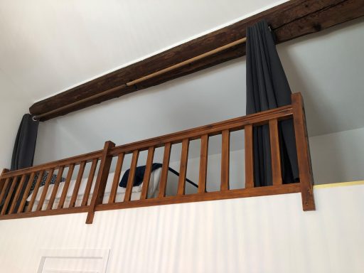 Balcon en bois avec barrières, rideaux gris et lit visible en haut.