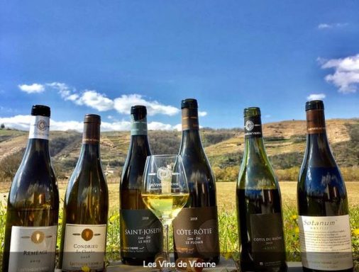 Six bouteilles de vin alignées avec un verre de vin blanc devant un paysage de collines.